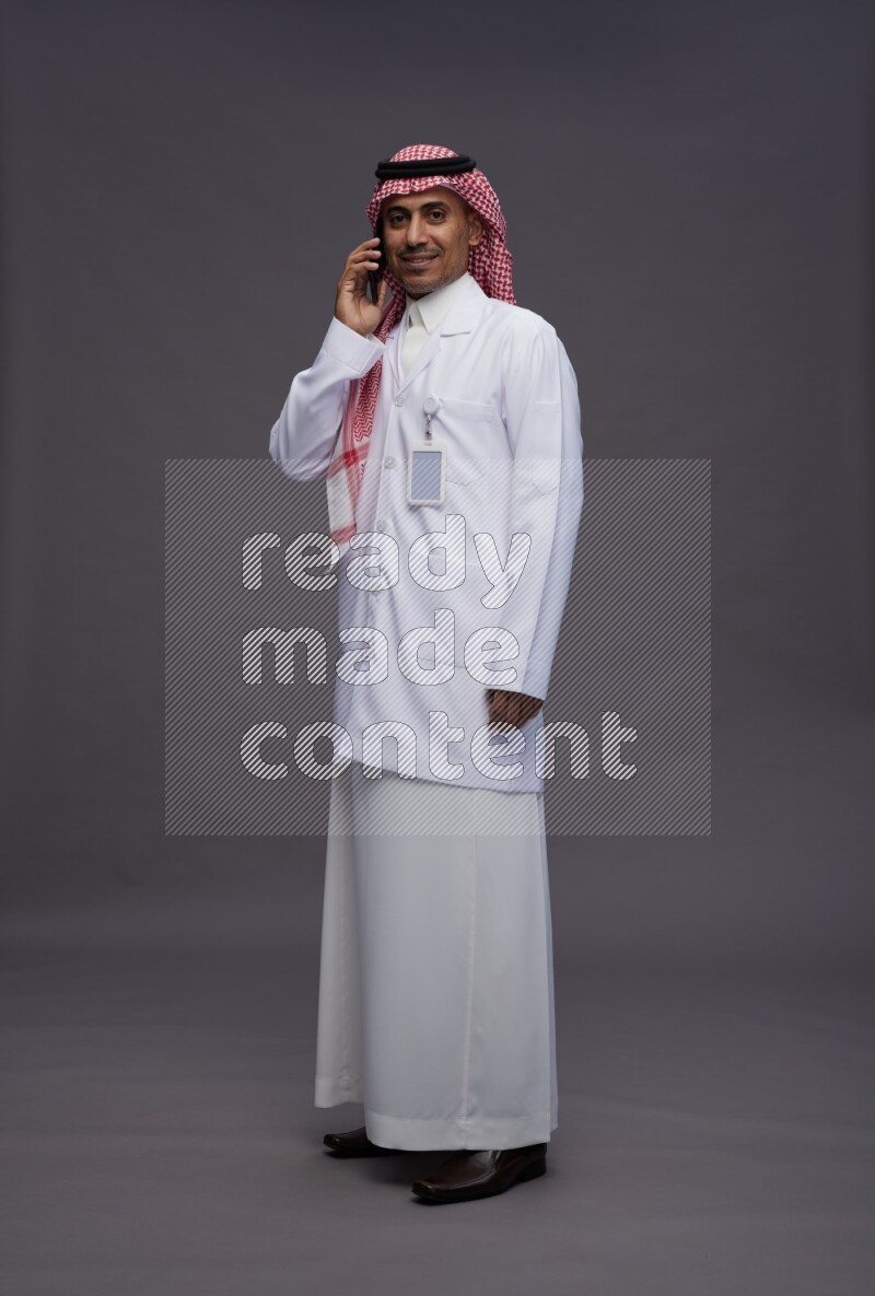 Saudi man wearing thob with lab coat and shomag with pocket employee badge standing talking on phone on gray background