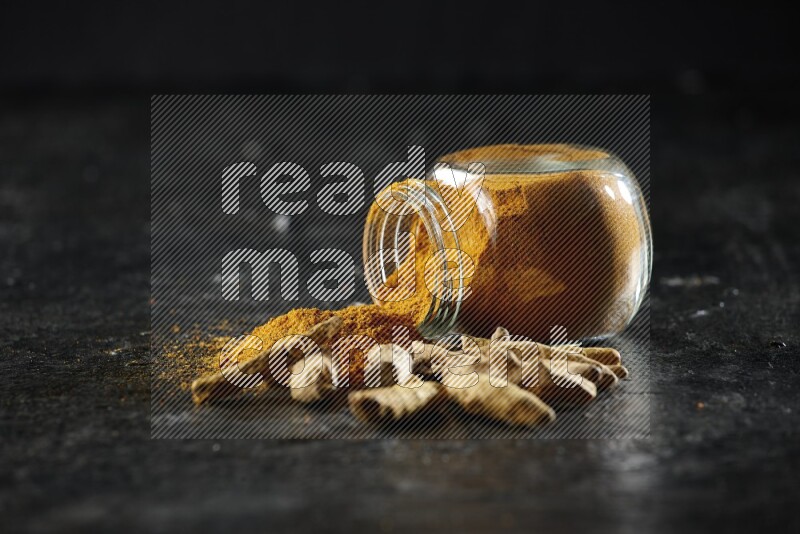 وعاء زجاجي للتوابل ممتلئ ببودرة الكركم مقلوب والبودرة تناثرت منه مع أصابع الكركم المجففة على خلفية سوداء