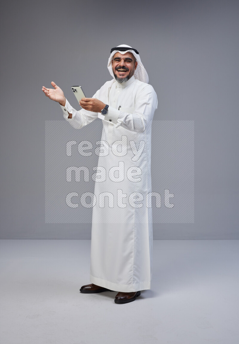 Saudi man Wearing Thob and white Shomag standing texting on phone on Gray background
