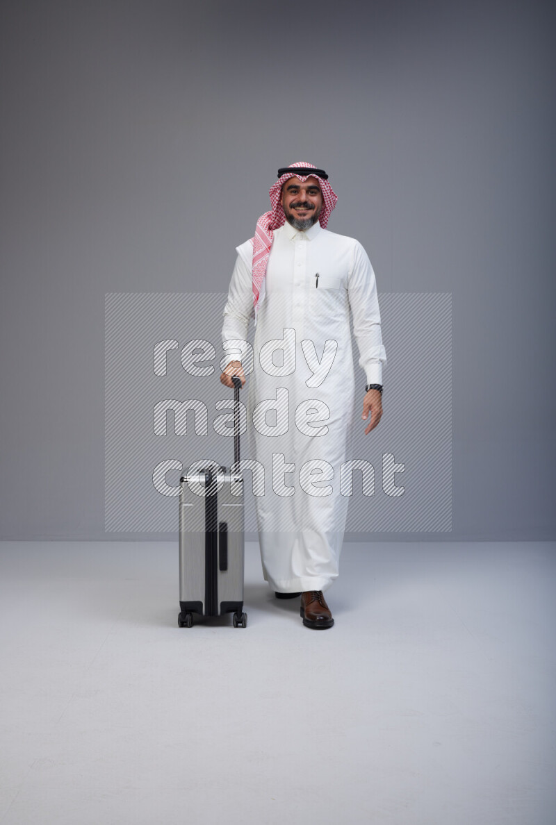 Saudi man wearing Thob and red Shomag standing holding Travel bag on Gray background