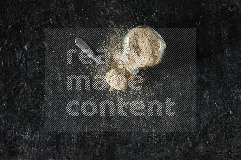 A flipped glass spice jar and metal spoon full of cardamom powder and powder spilled out of it on textured black flooring