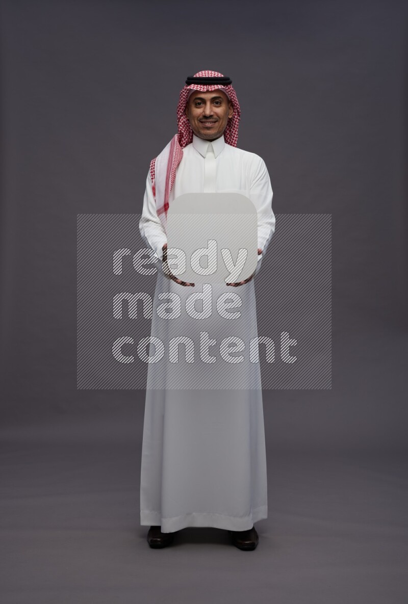 Saudi man wearing thob and shomag standing holding social media sign on gray background