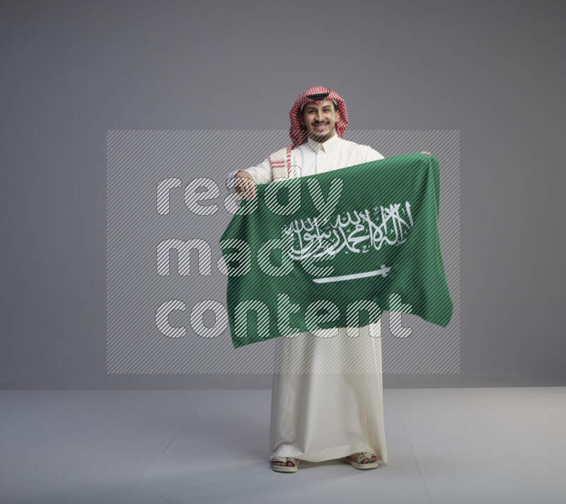 A saudi man standing wearing thob and red shomag holding big saudi flag on gray background