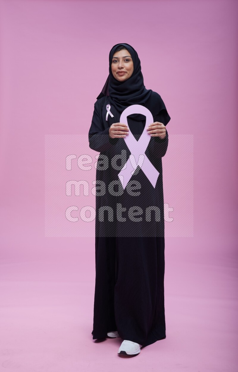 Saudi woman wearing pink ribbon on abaya standing holding awareness ribbon on plain pink background