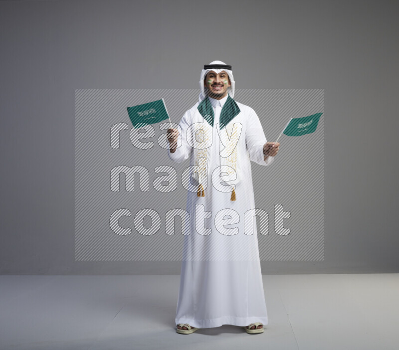 A saudi man standing wearing thob and white shomag with face painting and saudi flag scarf and holding small saudi flag on gray background