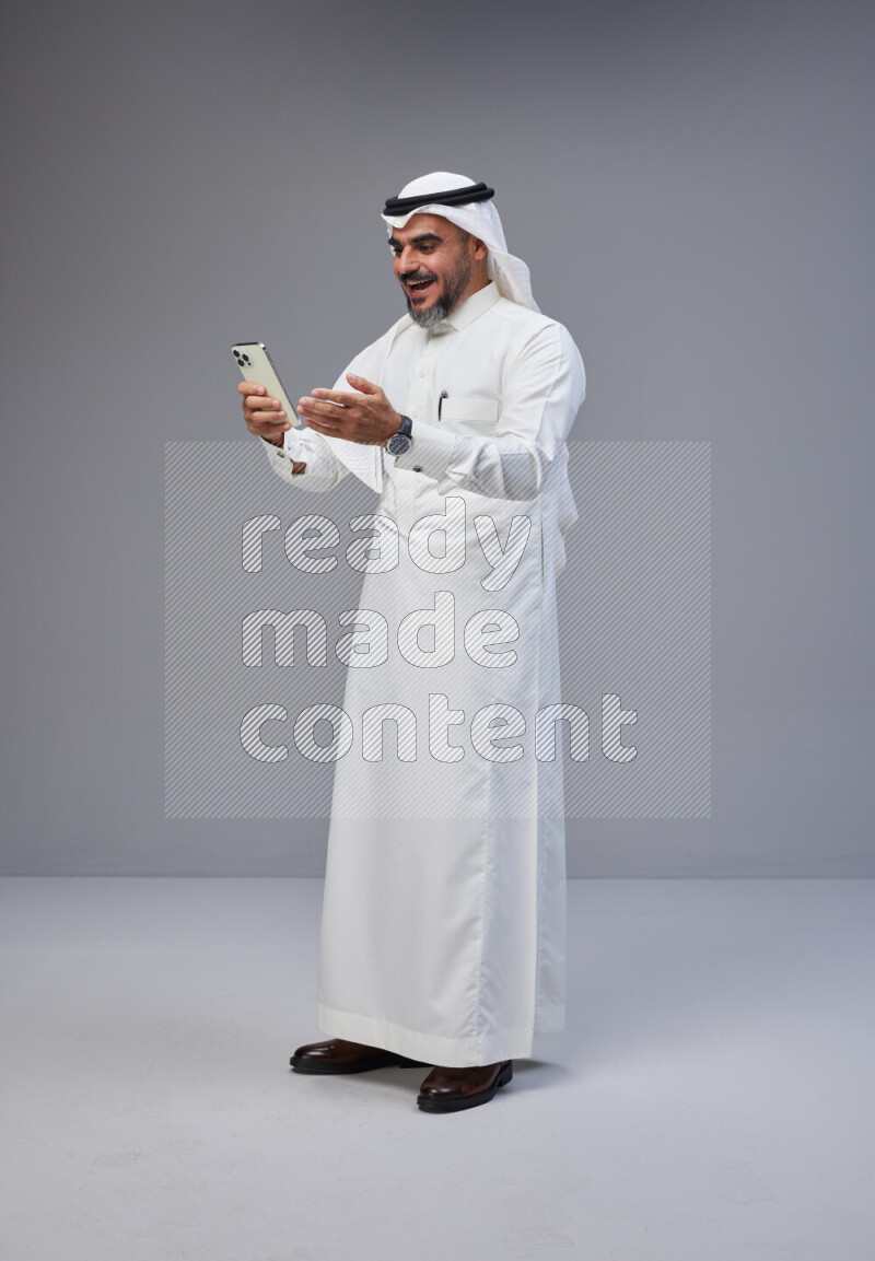 Saudi man Wearing Thob and white Shomag standing texting on phone on Gray background