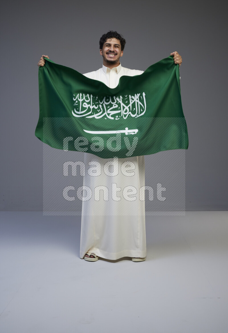 A saudi man standing wearing thob holding big saudi flag on gray background