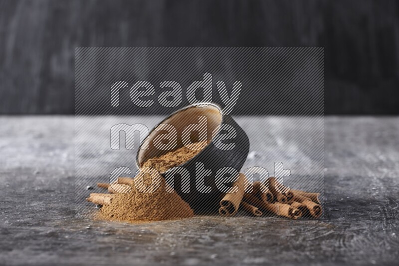 Black pottery bowl over filled with cinnamon powder and cinnamon sticks around the bowl on a textured black background