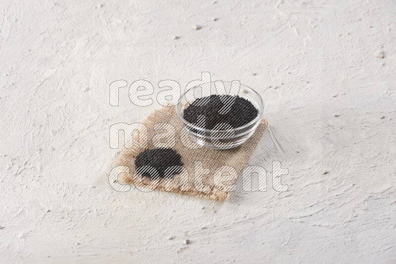 A glass bowl full of black seeds on a burlap piece on textured white flooring
