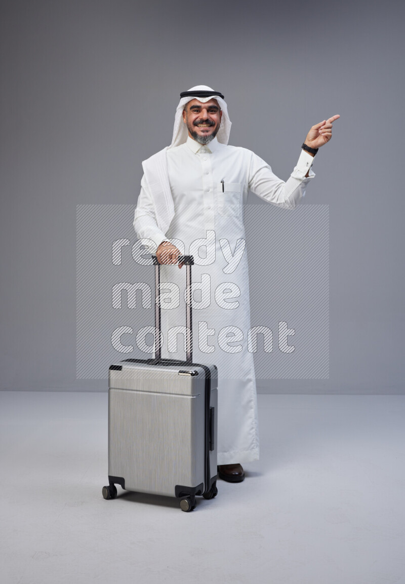 Saudi man wearing Thob and white Shomag standing holding Travel bag on Gray background