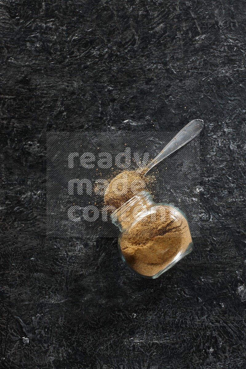A flipped glass spice jar and a metal spoon full of cumin powder and powder spilled out on a textured black flooring