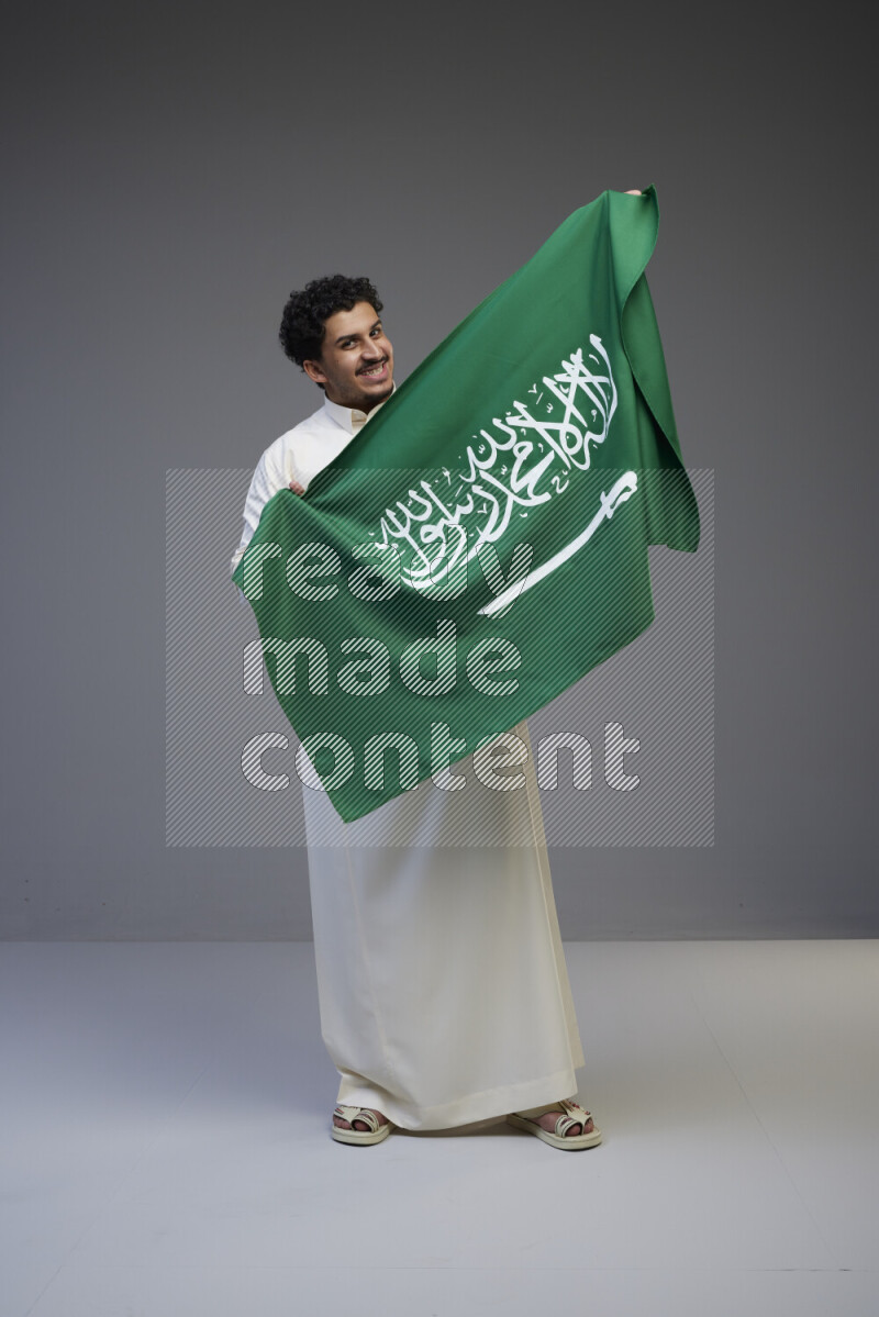 A Saudi man standing wearing thob holding big Saudi flag on gray background