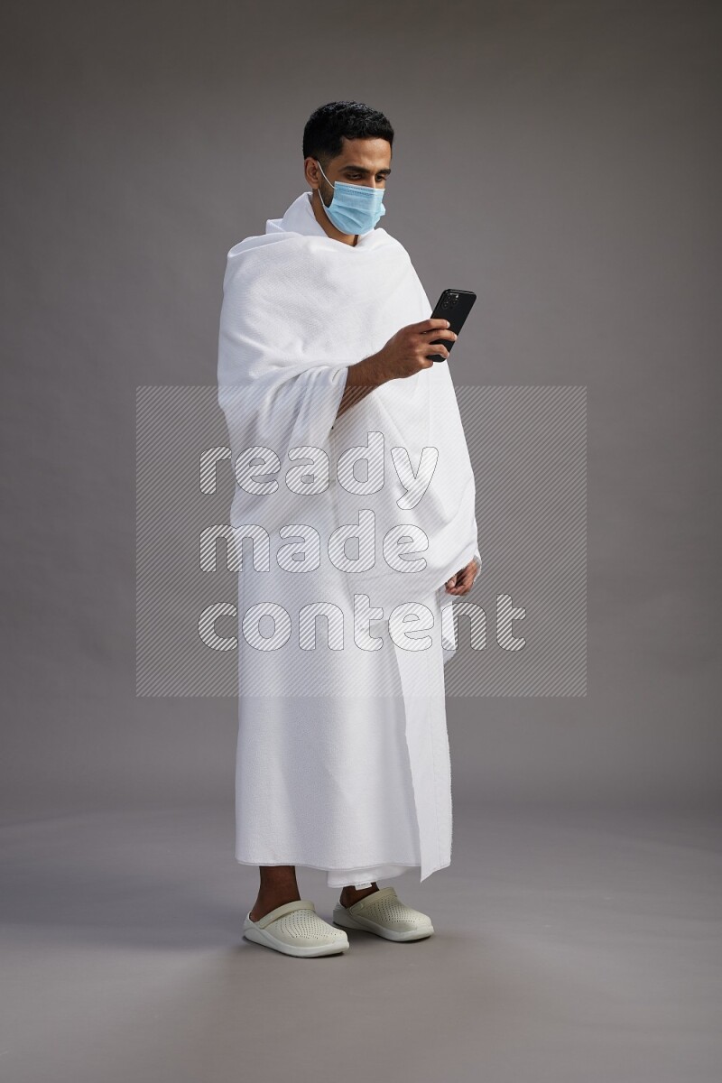 A man wearing Ehram with face mask Standing texting on phone on gray background