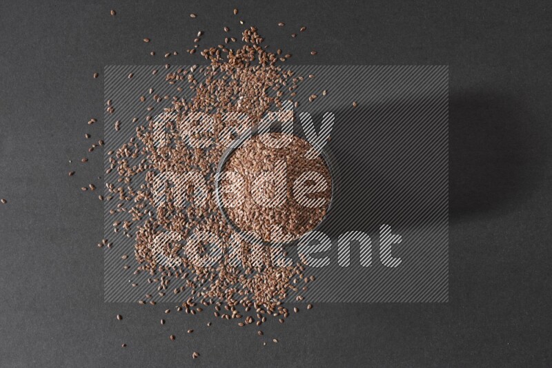 A black pottery bowl full of flaxseeds surrounded by the seeds on a black flooring