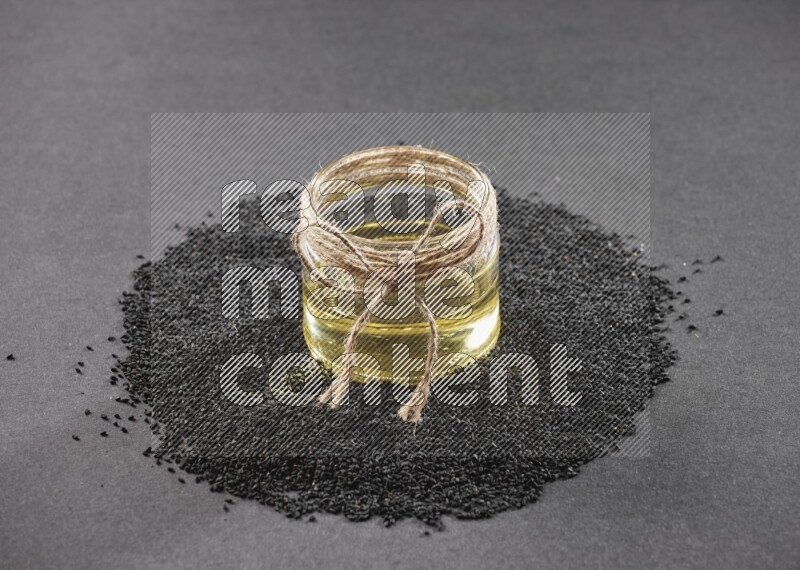 A glass jar full of black seeds oil surrounded by the seeds on a black flooring