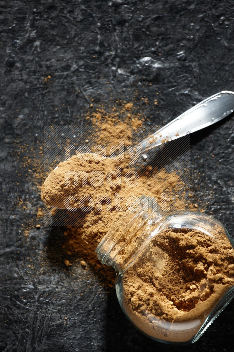 A flipped glass spice jar and a metal spoon full of allspice powder and powder spilled out of it on a textured black flooring
