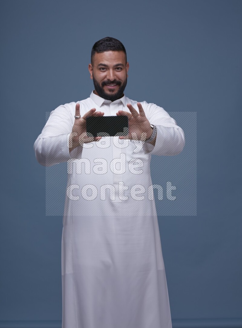 A man Showing his phone in Blue Background wearing Saudi Thob