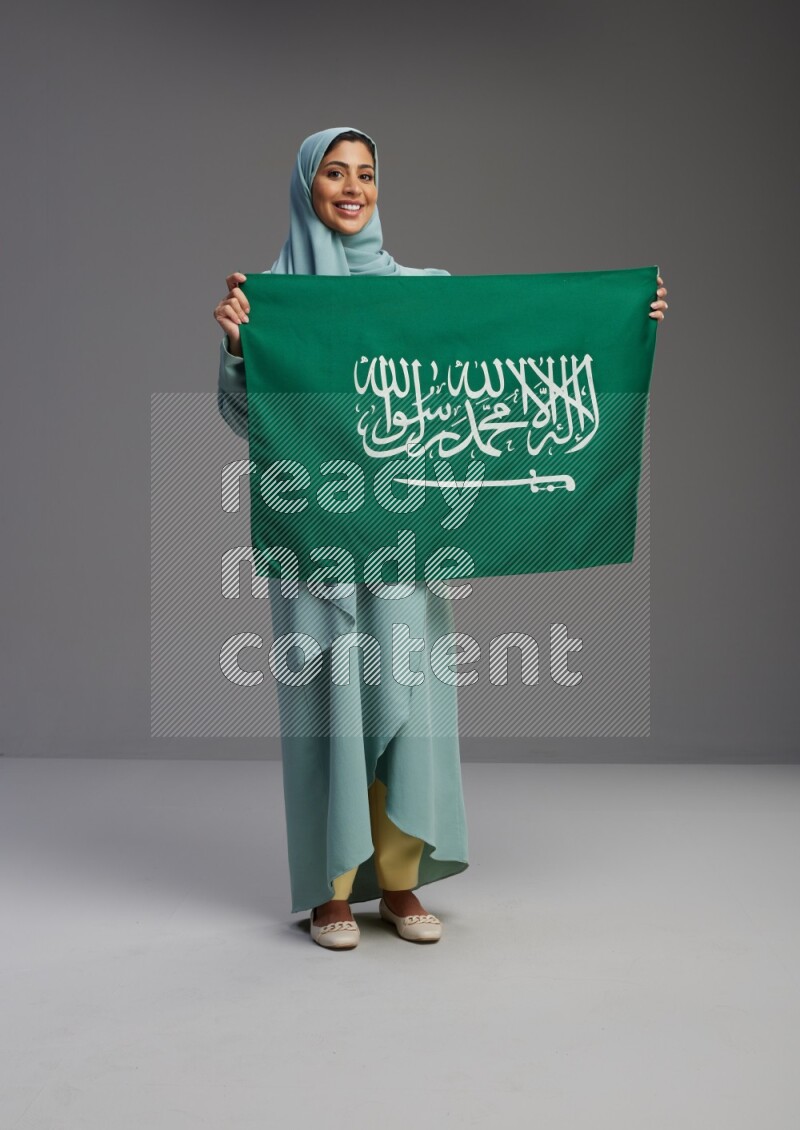 Saudi Woman wearing Abaya standing holding Saudi flag on Gray background