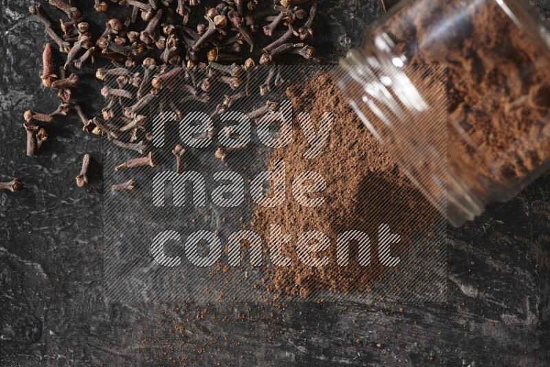 A flipped glass jar full of cloves powder with cloves spread on a textured black flooring