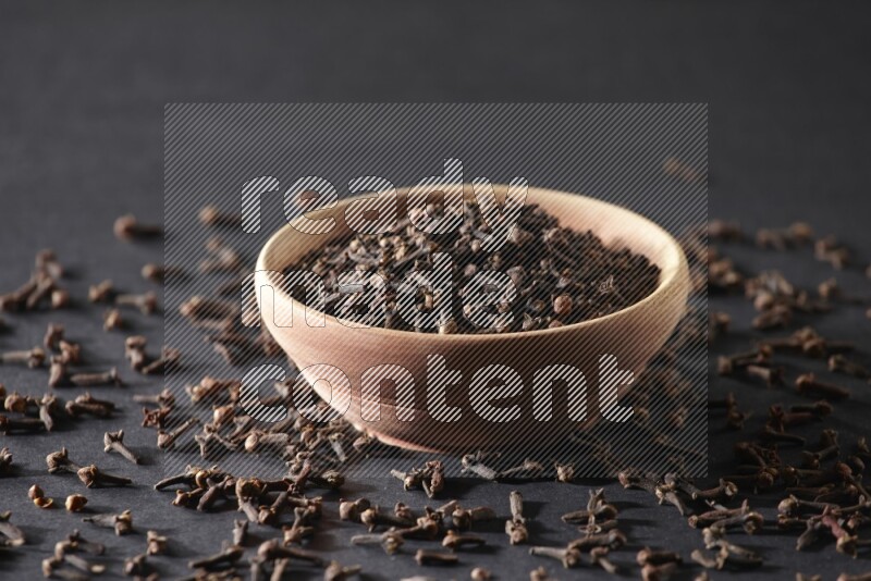 A wooden bowl full of cloves with spreaded whole cloves on a black flooring