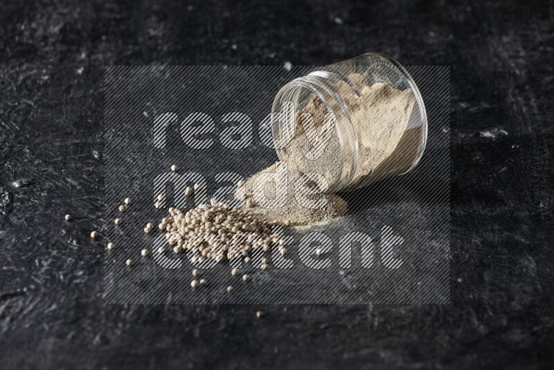 A flipped glass jar full of white pepper powder with spilled powder and pepper beads on textured black flooring
