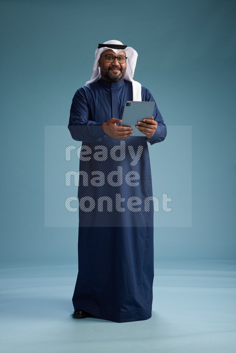 Saudi Man with shimag Standing holding ATM card while working on tablet on blue background