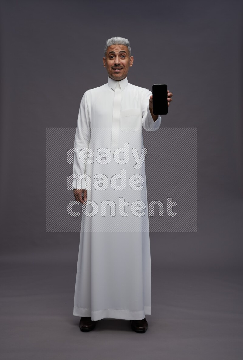 Saudi man wearing thob standing showing phone to camera on gray background