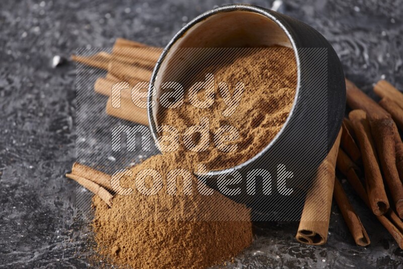 Black pottery bowl over filled with cinnamon powder and cinnamon sticks around the bowl on a textured black background