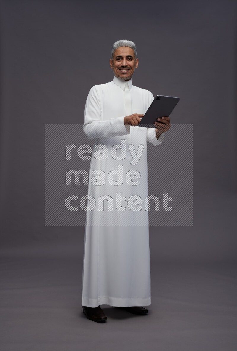 Saudi man wearing thob standing working on tablet on gray background