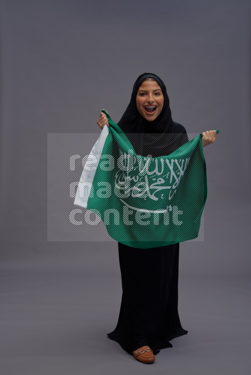 Saudi woman wearing Abaya standing holding Saudi flag on gray background