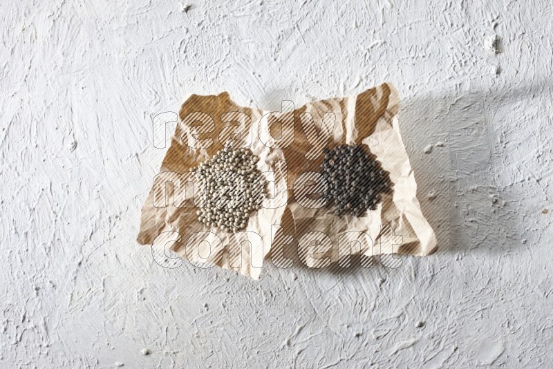 2 Crumpled pieces of paper full of black and white pepper beads on a textured white flooring