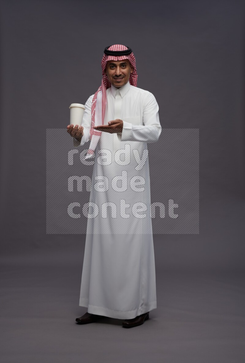 Saudi man wearing thob and shomag standing holding paper cup on gray background