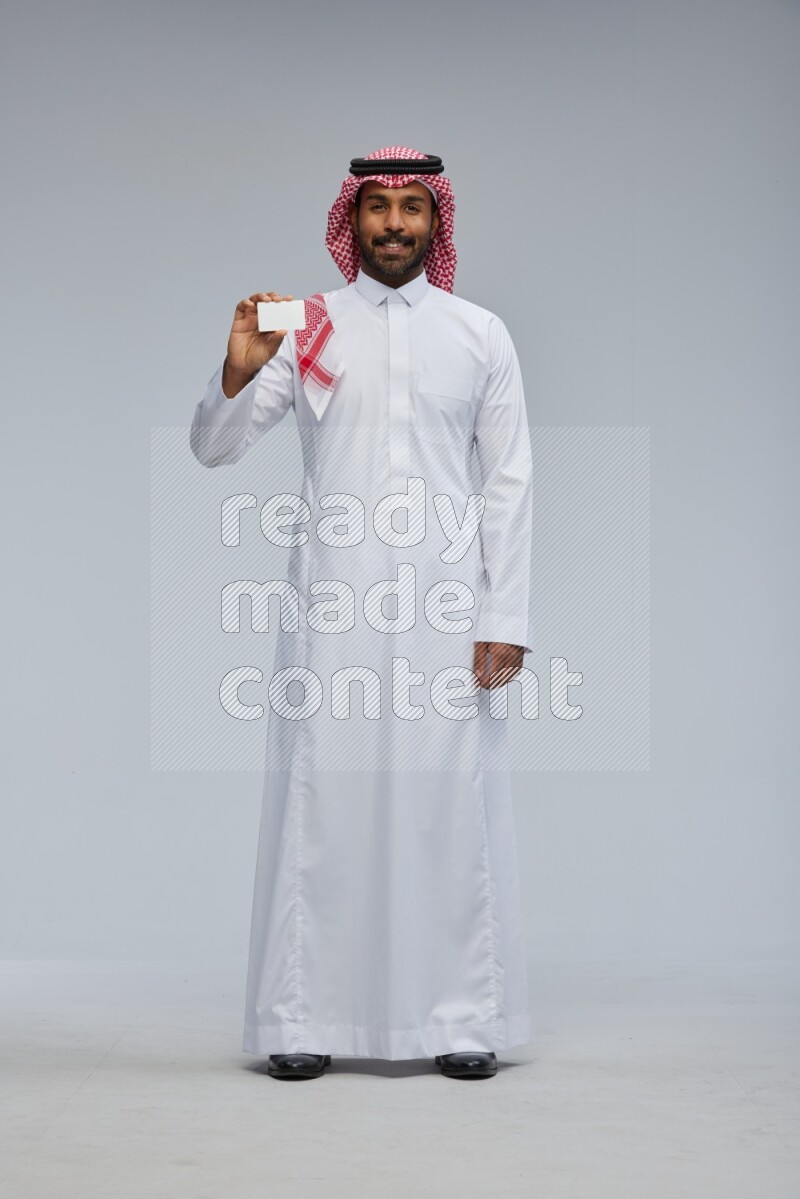 Saudi man wearing thob and shomag standing holding ATM card on gray background
