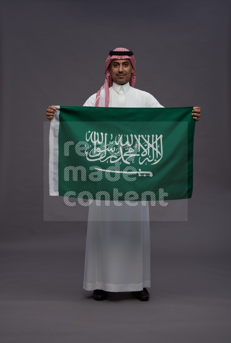 Saudi man wearing thob and shomag standing holding Saudi flag on gray background