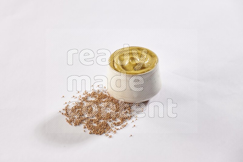 A beige pottery bowl full of mustard paste with mustard seeds underneath on white flooring