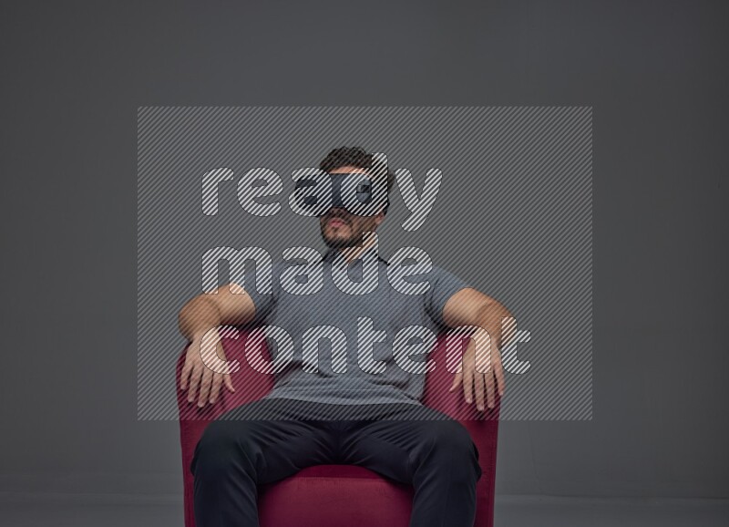 A man wearing casual and wearing VR while sitting on a burgundy chair eye level on a gray background