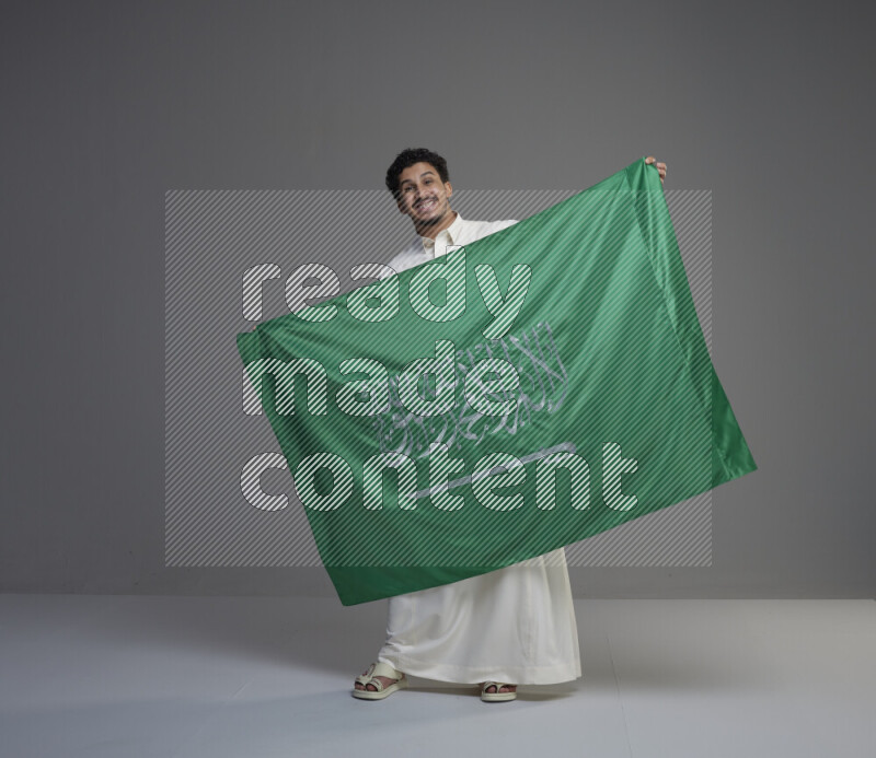 A Saudi man standing wearing thob holding big Saudi flag on gray background
