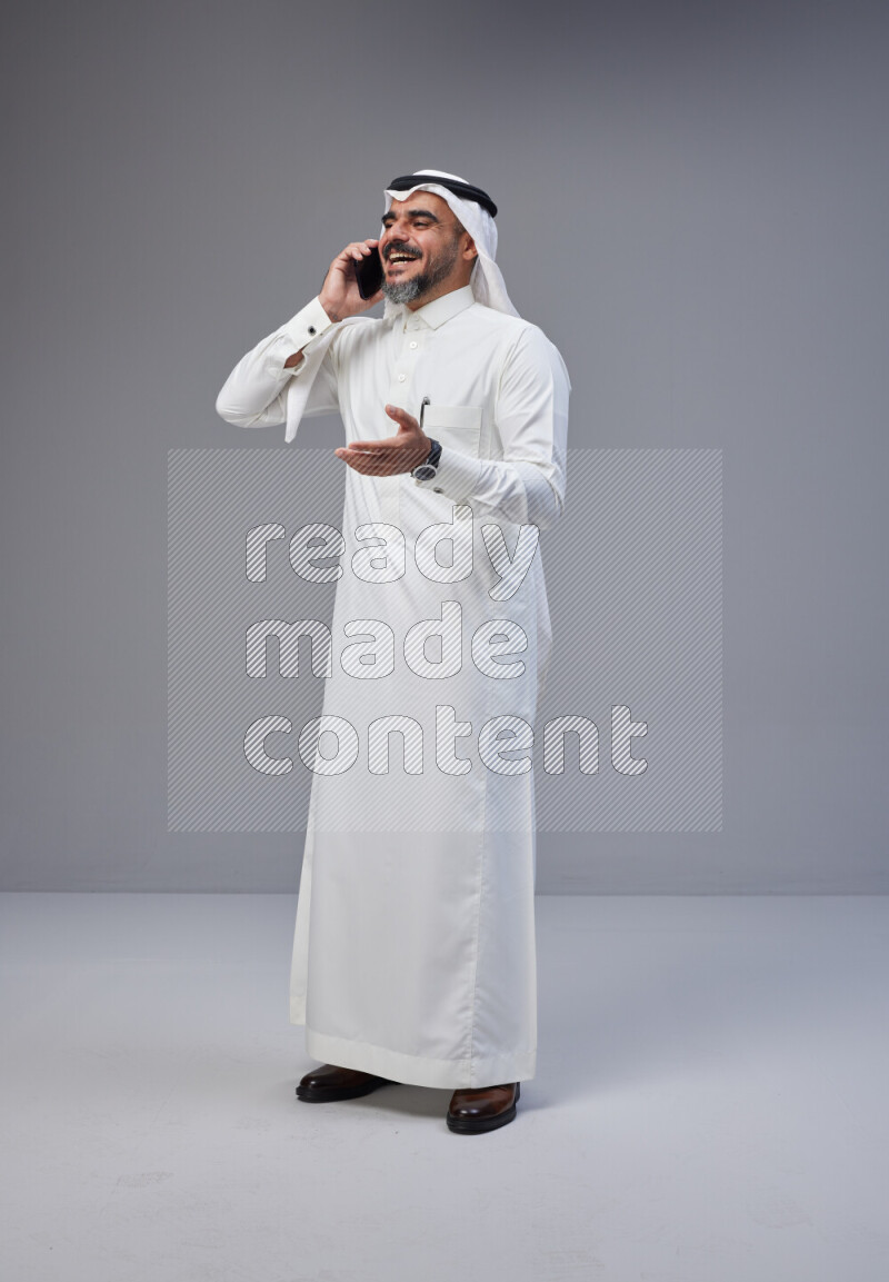 Saudi man Wearing Thob and white Shomag standing talking on phone on Gray background