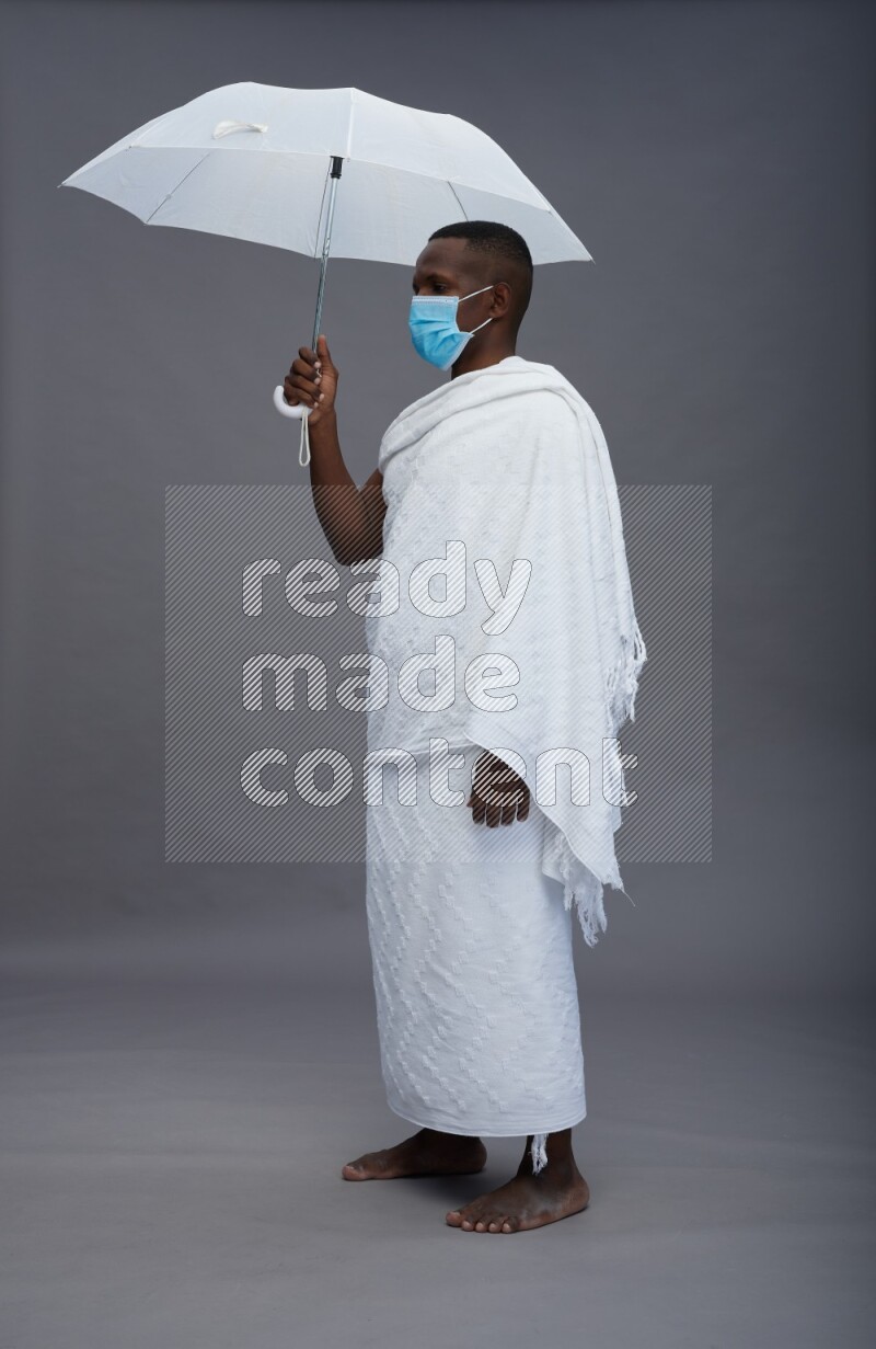 A man wearing Ehram with face mask Standing holding umbrella on gray background
