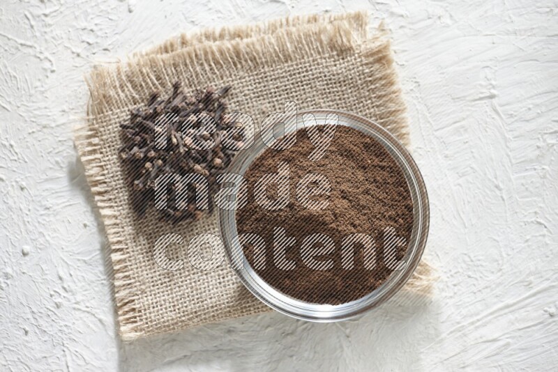 A glass bowl full of cloves powder and cloves grains on a burlap piece on a white flooring