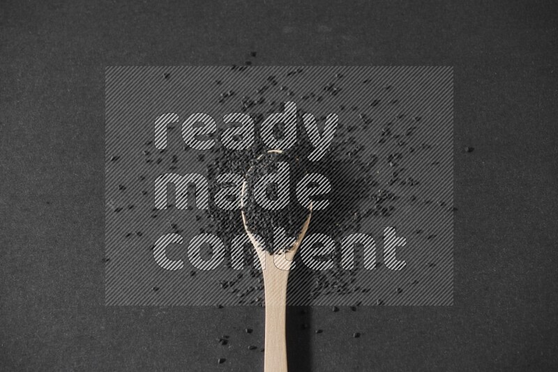 A wooden spoon full of black seeds and surrounded by seeds on a black flooring