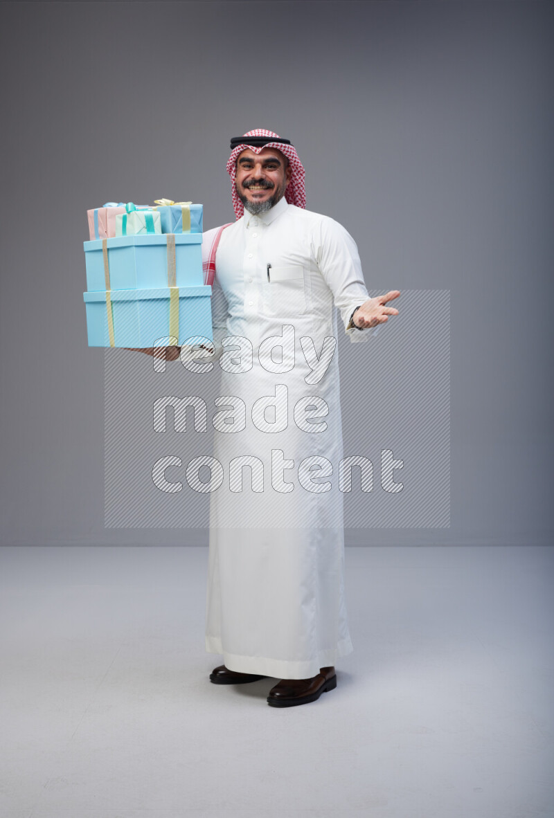 Saudi man Wearing Thob and red Shomag standing holding gift box on Gray background