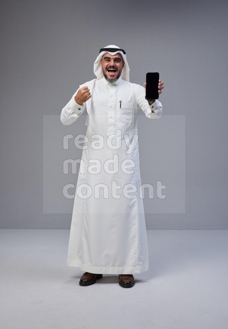 Saudi man Wearing Thob and white Shomag standing showing phone to camera on Gray background