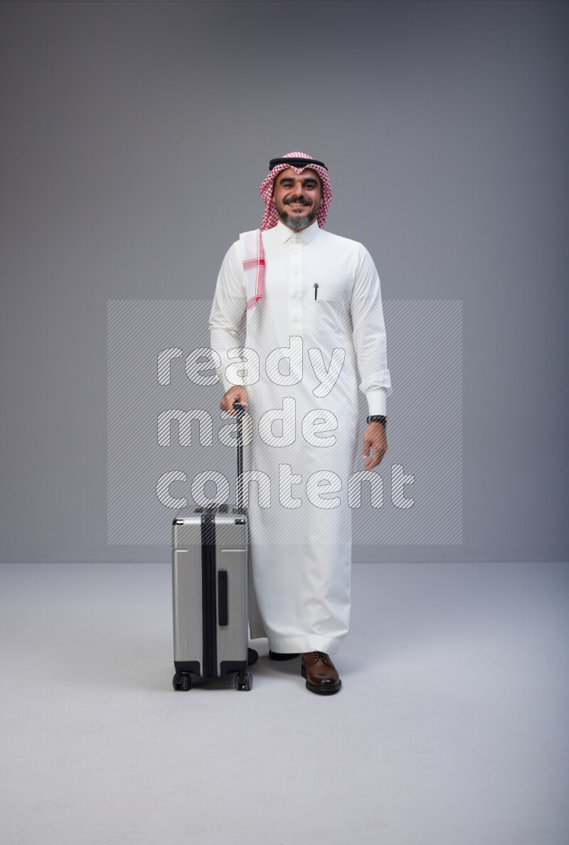Saudi man wearing Thob and red Shomag standing holding Travel bag on Gray background