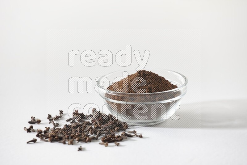 A glass bowl full of cloves powder and cloves grains spread on white flooring