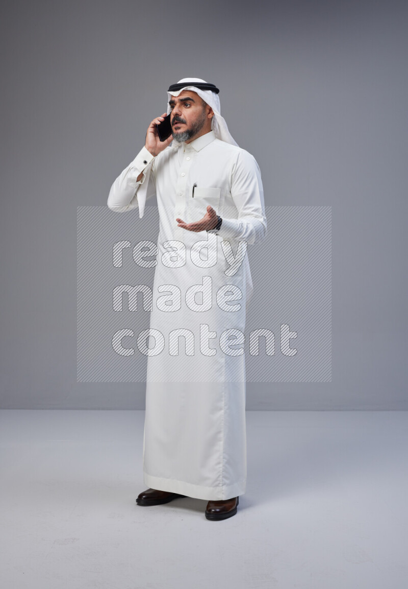 Saudi man Wearing Thob and white Shomag standing talking on phone on Gray background