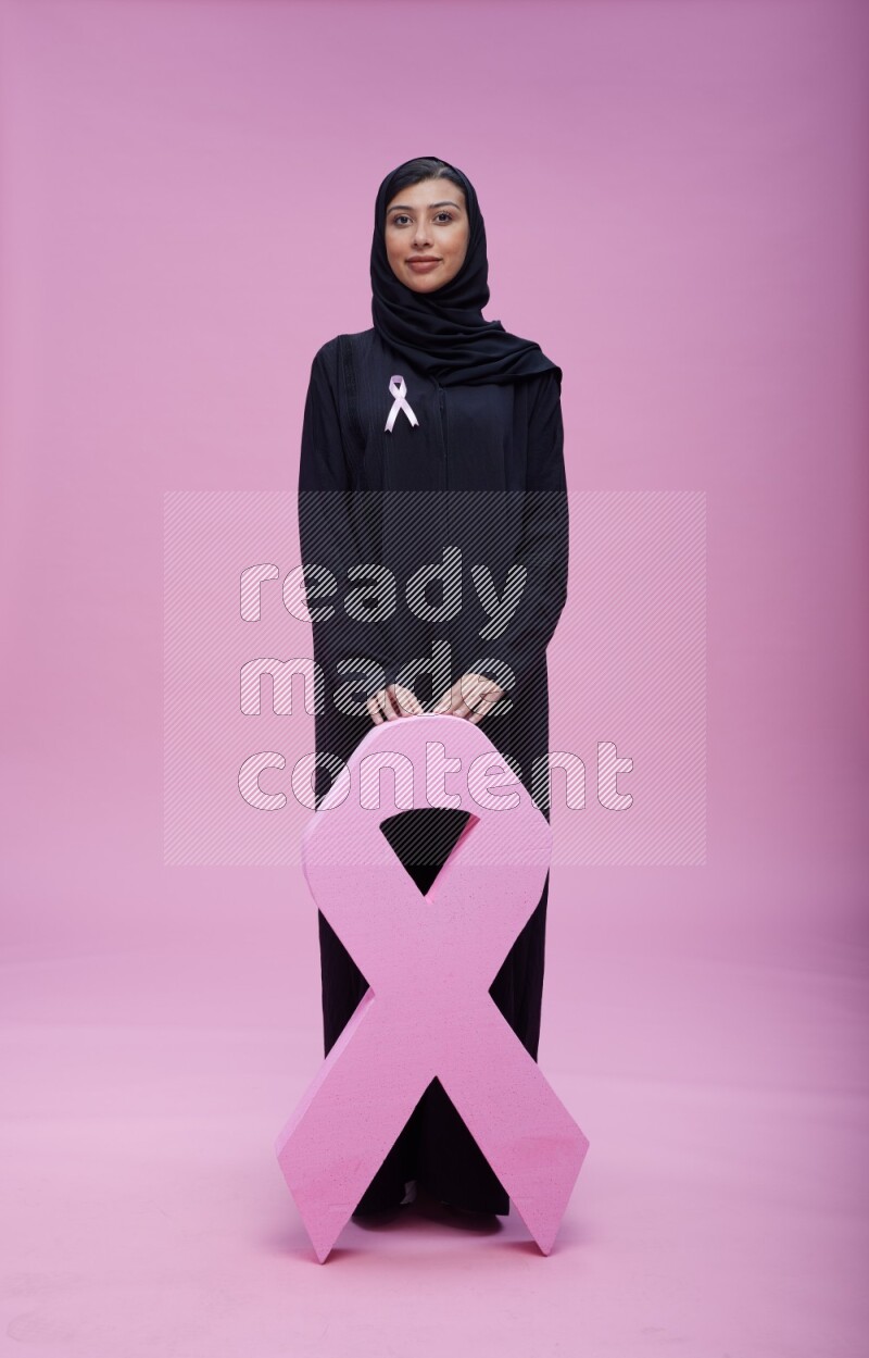 Saudi woman wearing pink ribbon on abaya standing holding awareness ribbon on plain pink background