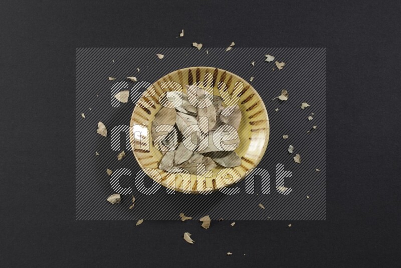 A multicolored pottery plate filled with dried bay leaves on black flooring