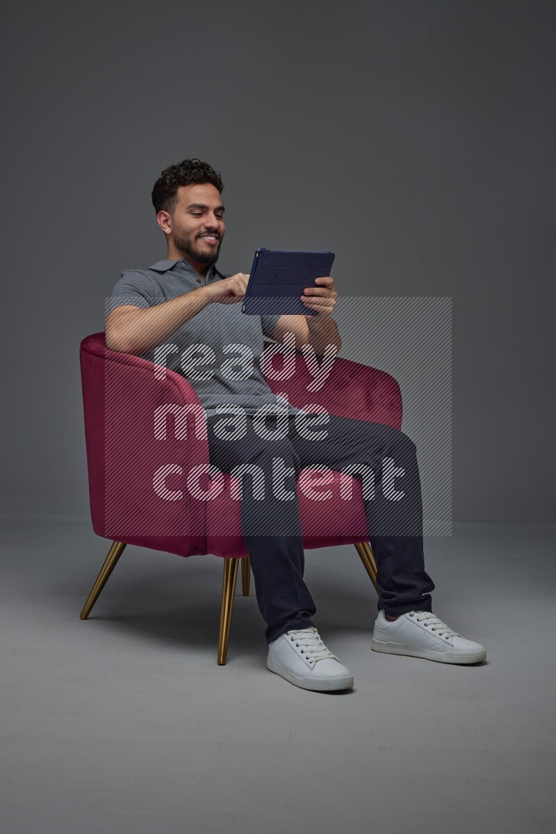 A man wearing casual and using his tablet while sitting on a burgundy chair eye level on a gray background
