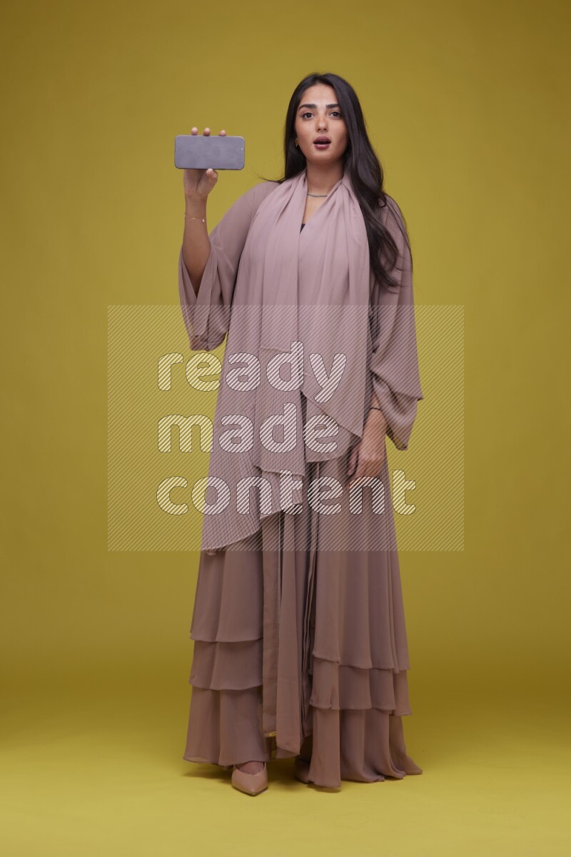 A woman Showing SmartPhone Screen on a Yellow Background wearing Brown Abaya